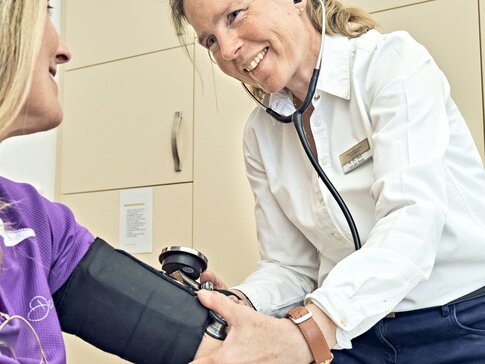 Ich sehe eine Ärztin mit weißer Bluse und dunkelblauer Hose, die eine blonde Patientin in lila T-Shirt mit Stethoskop und Blutdruckmanschette untersucht.