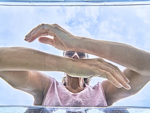 Hier sehen Sie eine Frau mit pinkem Shirt, welche ihre Arme in klares Wasser taucht. Die Aufnahme wurde aus dem Wasser, unterhalb der Frau, aufgenommen.