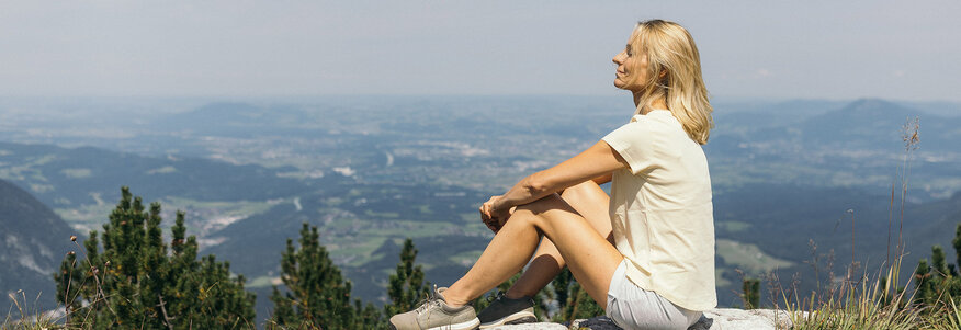Frau sitzt auf einem Stein in der Sonne, im Hintergrund Landschaftspanorama.