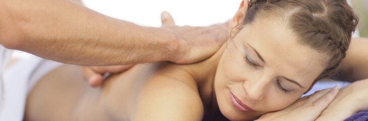 Hier sehen Sie eine Frau, die mit geschlossenen Augen auf einer Massageliege liegt. Sie liegt mit dem Bauch nach unten und Männerhände massieren ihr den oberen Rücken. 