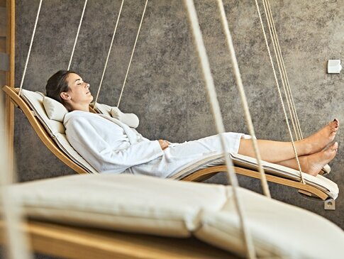 Hier sehen Sie mehrere Hängeliegen in einem Entspannungsraum. Auf der hintersten Liege liegt eine Frau in einem weißen Bademantel. 