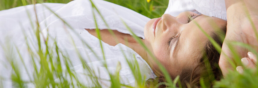 Hier sehen Sie eine Frau, welche auf einer Wiese liegt. Neben ihr liegt ein Buch. Sie hat die Augengeschlossen und lächelt. 