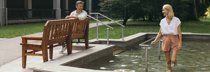 Frau kneippt und redet mit ihrem Mann, der auf der Bank sitzt