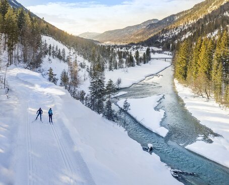 Hier sehen Sie eine Schneelandschaft entlag eines Flusses in Mitten der Berge. Zwei Personen sieht man in der Ferne. 