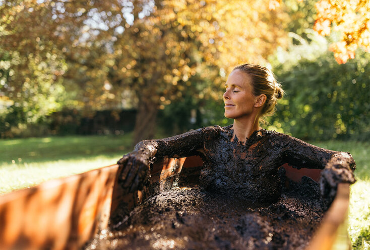 Frau sitzt mit Moor bedeckt bei einer Kur in einer hölzernen Badewanne draußen in der Herbstsonne