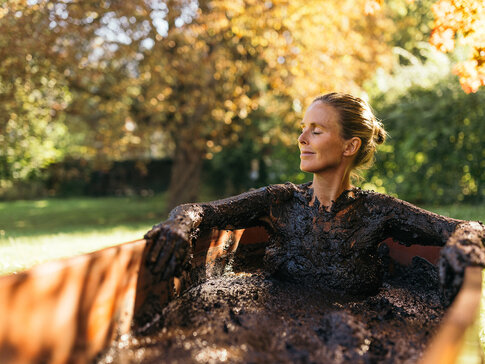 Frau sitzt mit Moor bedeckt bei einer Kur in einer hölzernen Badewanne draußen in der Herbstsonne