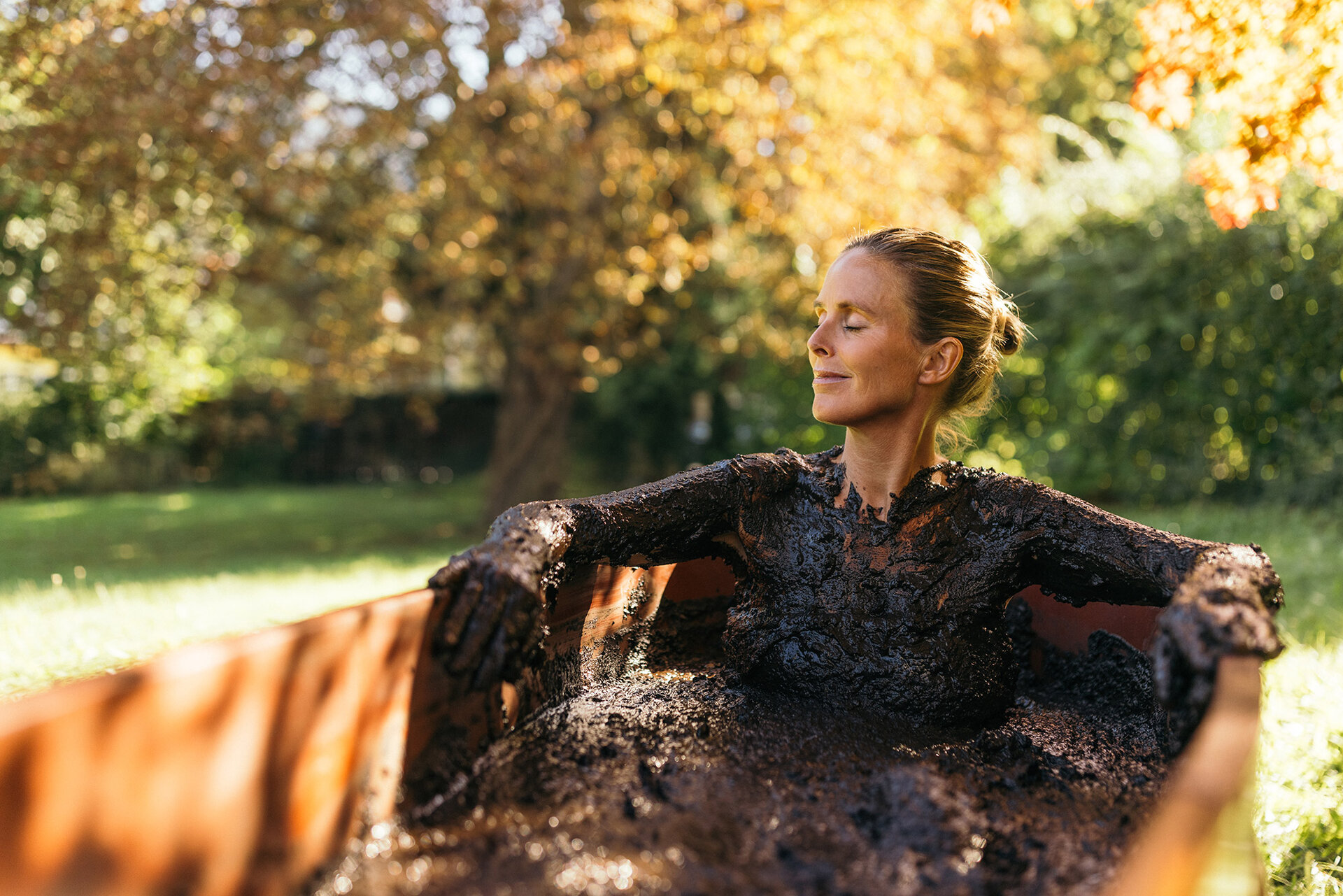 Frau sitzt mit Moor bedeckt bei einer Kur in einer hölzernen Badewanne draußen in der Herbstsonne