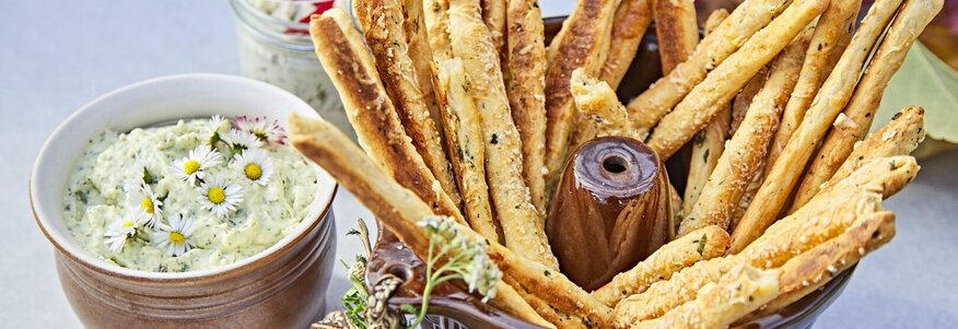 Hier sehen Sie auf einem Tisch eine kleine Schüssel mit selbstgemachtem frischen Kräuterquark. In einer größeren Schüssel daneben sind frische Teigstangen. Dekoriert ist der Tiasch mit Kräutern und Blüten.