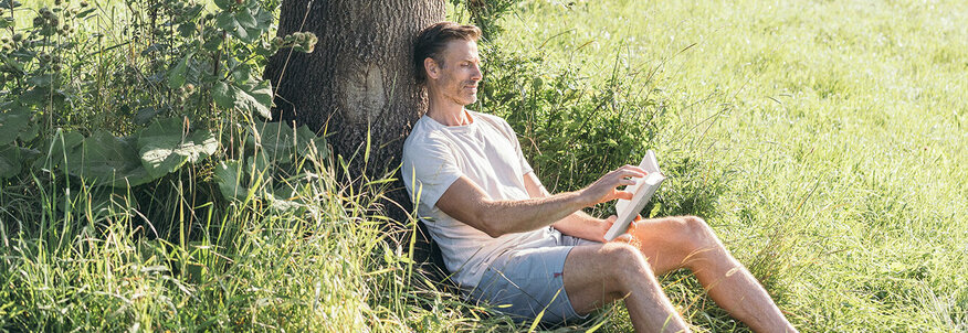 Der Mann sitzt angelehnt an einem Baum mit einem Buch