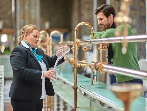 Hier sehen Sie eine Brunnenhalle in der Heilwasser aus goldenen Zapfhänen ausgeschenkt wird. Eine Frau in Einheitskleidung schenkt einem Mann der zu Gast ist gerade ein Glas Heilwasser ein. 