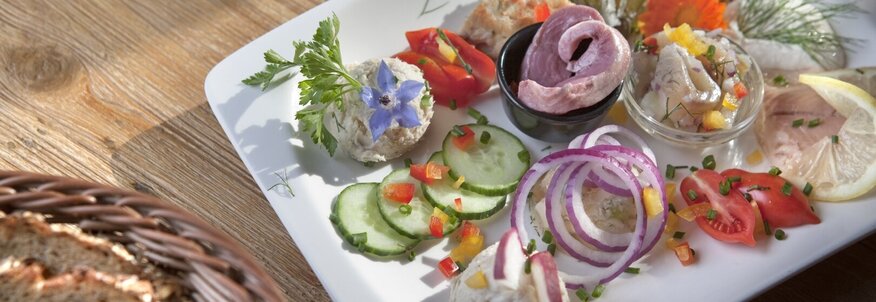 Hier sehen Sie einen Brotzeitteller mit viel Gemüse, wie Gurke und Tomate, ebenso sehen Sie unterschiedliche Brotaufstriche und dunkles Bauernbrot. 