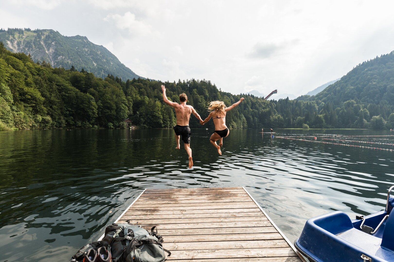 Eine Frau und ein Mann springen händehaltend von einem Steg in einen See. Neben dem Steg befindet sich ein Tretboot. Der See ist umgeben von Wald und Bergen.
