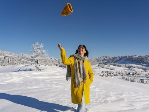 Hier sehen Sie eine Frau in gelbem Mantel, die in einer mit Schnee bedeckten Landschaft lachen ihre Mütze in die Luft wirft.