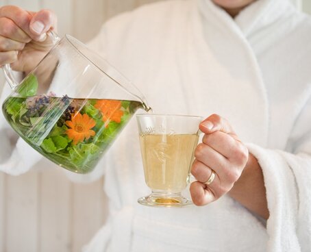 Hier sehen Sie eine Person im Badementel. In der einen Hand hat die Person einen Teetasse in der anderen Hand einen frisch aufgegossenen Tee mit frischen Kräutern. Die Personen schenkt den Tee in die Teetasse ein. 