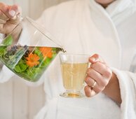 Hier sehen Sie eine Person im Badementel. In der einen Hand hat die Person einen Teetasse in der anderen Hand einen frisch aufgegossenen Tee mit frischen Kräutern. Die Personen schenkt den Tee in die Teetasse ein. 