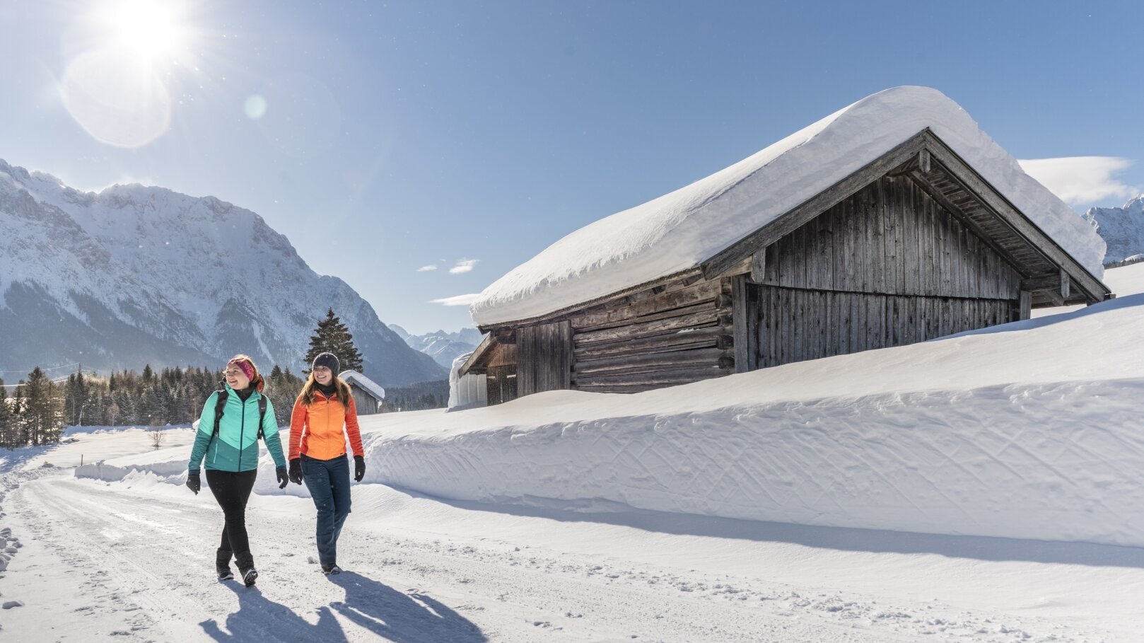 Hier sehen Sie zwei Personen in einer Winterlandschaft wandern. Sie gehen auf einer geräumten Straße, neben ihnen ist der Schnee noch sehr hoch. Sie gehen an einer kleinen Hütte vorbei und die Sonne scheint. 