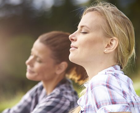 Nahaufnahme von zwei Frauen, die in einer Wiese sitzen und tief durchatmen.