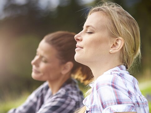 Nahaufnahme von zwei Frauen, die in einer Wiese sitzen und tief durchatmen.