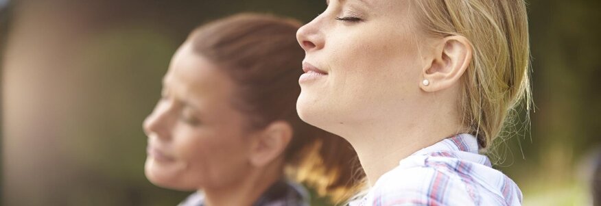 Nahaufnahme von zwei Frauen, die in einer Wiese sitzen und tief durchatmen.